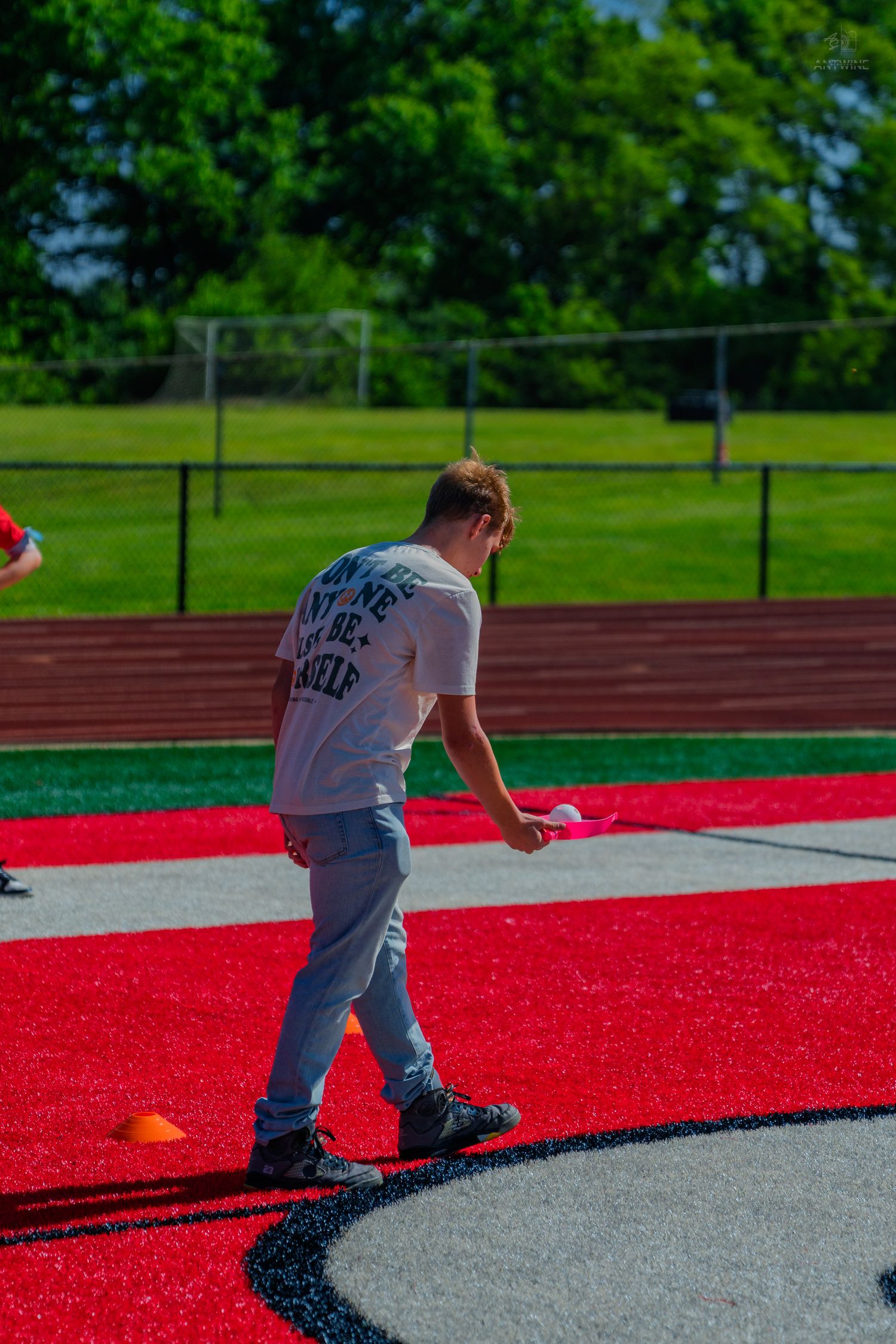 Young man in white t-shirt and gray pants walking on red turf field during outdoor sports activity.