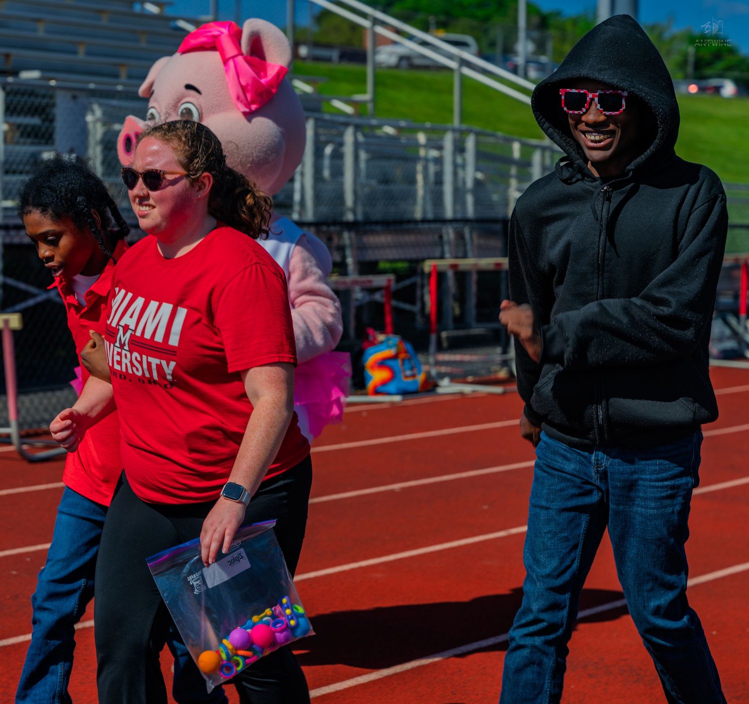 People walking on a red track at a university event with a pink pig mascot costume in the background.