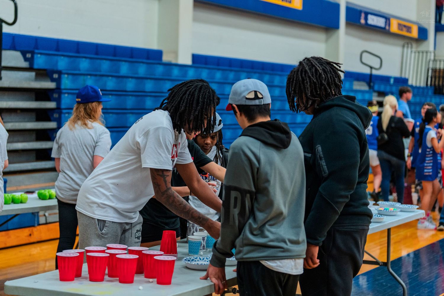 Students gather around a table with red Solo cups at an indoor school gymnasium event or party.