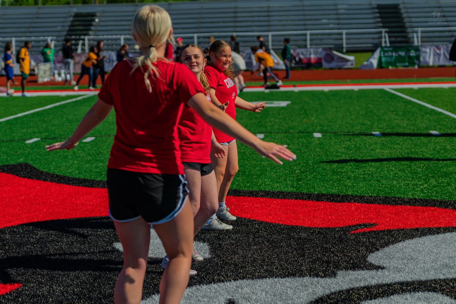 Girls in red shirts practice drills on a turf football field during an outdoor sports training session.