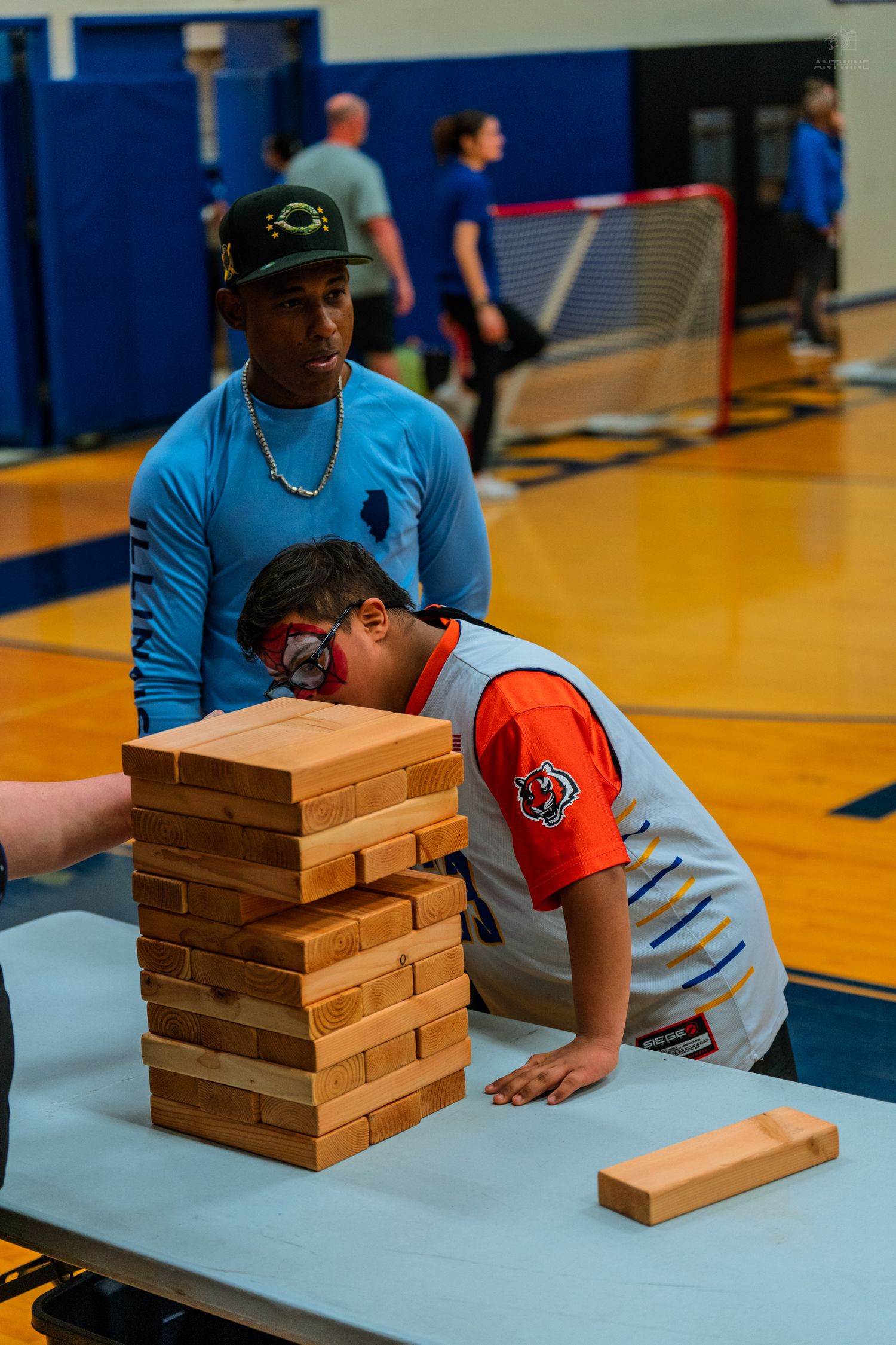 Child in Spider-Man mask carefully removes a block from a giant Jenga tower during an indoor gym activity event.