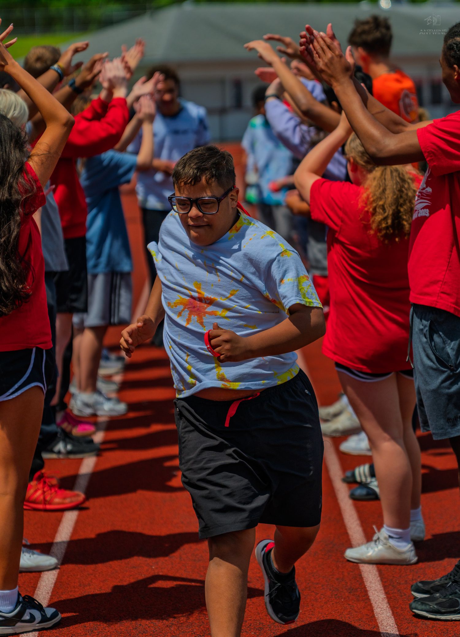 A young athlete runs through a cheering crowd on a red track during a Special Olympics event on a sunny day.