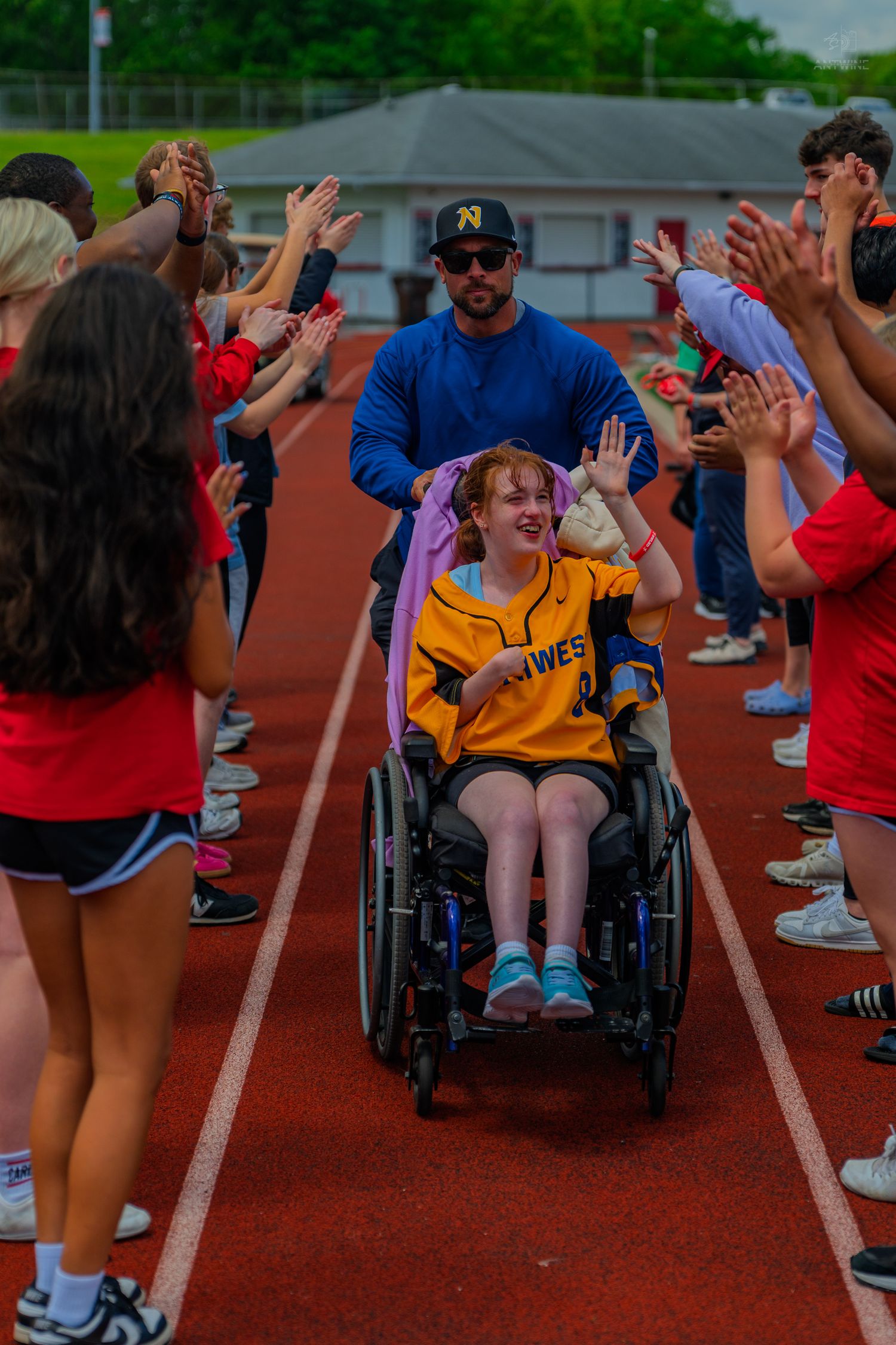 Girl in wheelchair high-fived by cheering students on a red track during a celebratory school event.