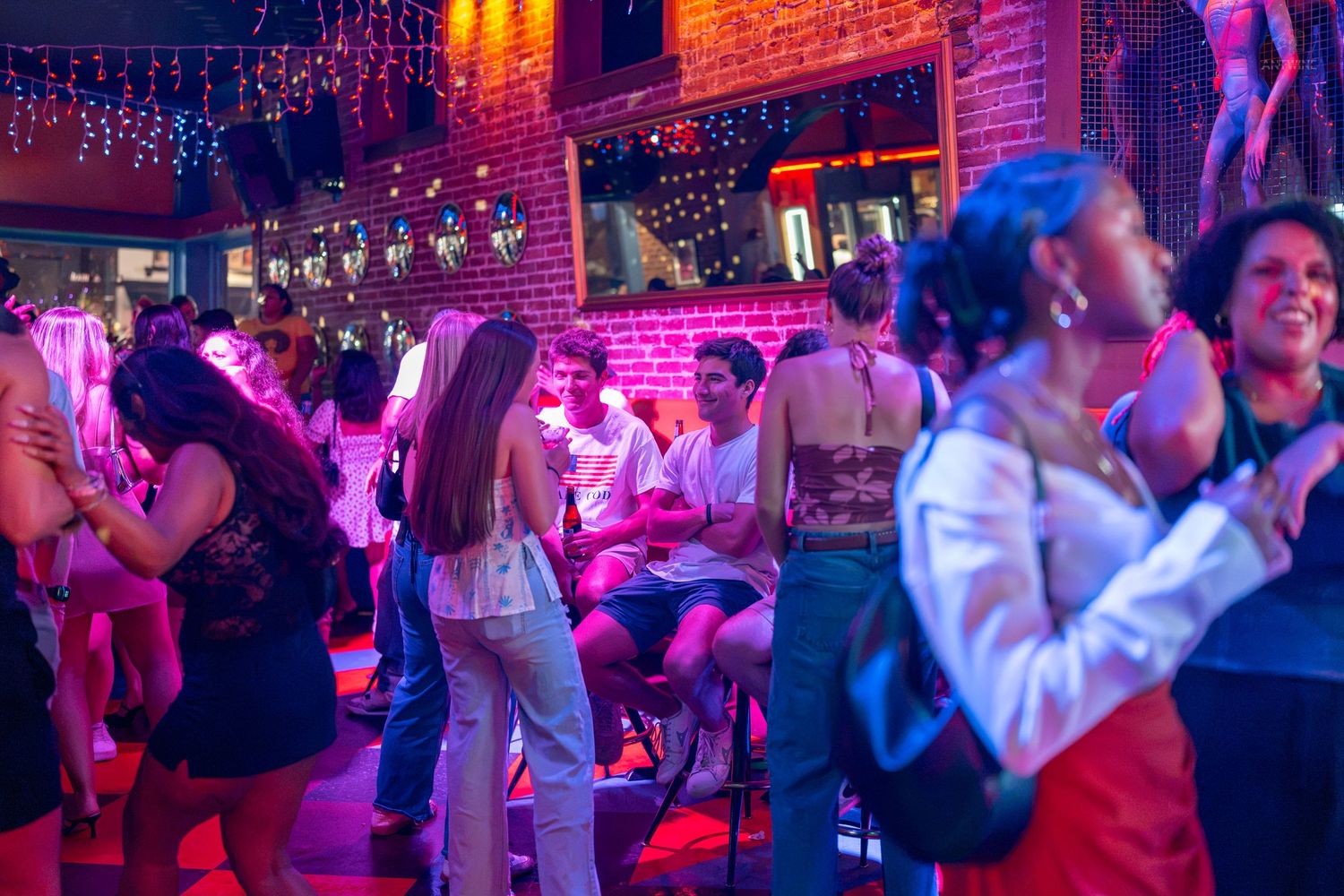 Crowded nightclub with people dancing under vibrant pink and purple lights in a brick-walled venue.