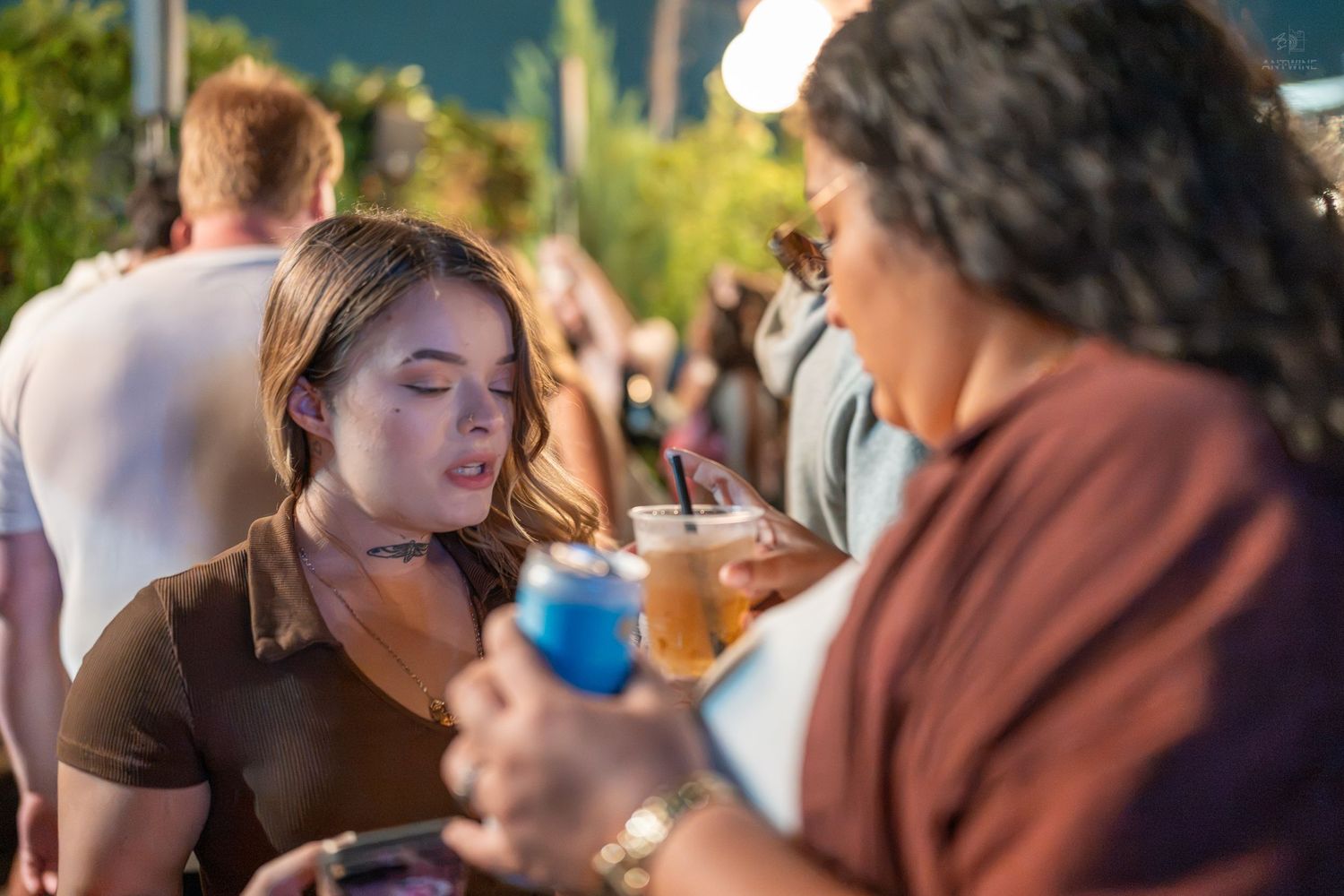 Two women socializing at an outdoor festival, holding colorful drinks in a lively crowd setting.
