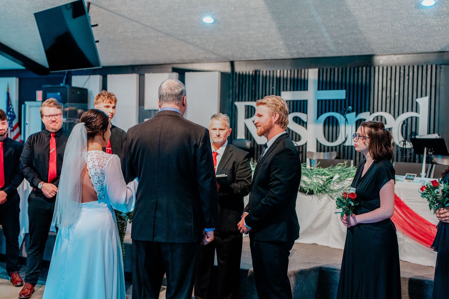 Bride and groom exchange vows at indoor wedding ceremony with guests and officiant gathered around.