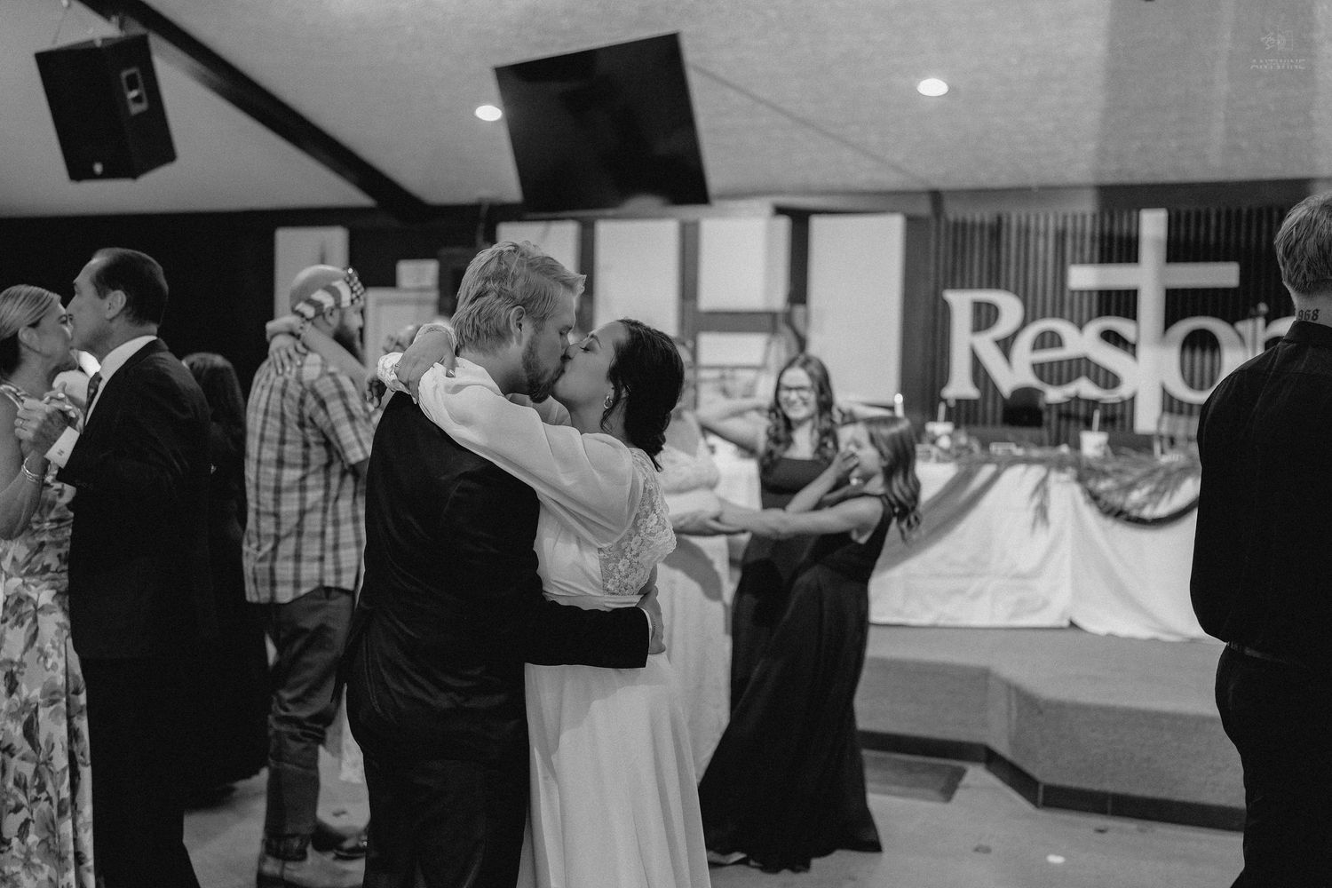 Couples dancing at wedding reception inside church hall with Restore sign in background.