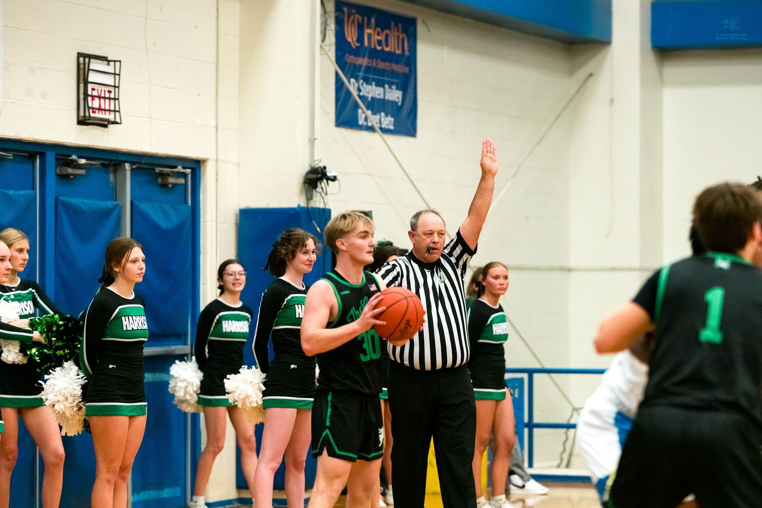 Referee signals while green uniformed basketball player holds ball near cheerleaders in gym.
