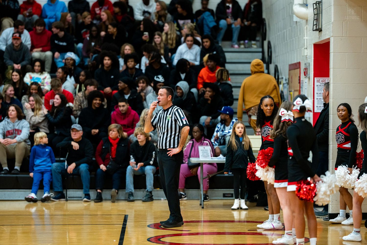 Basketball referee signals call on court while cheerleaders and large crowd watch high school gymnasium game.