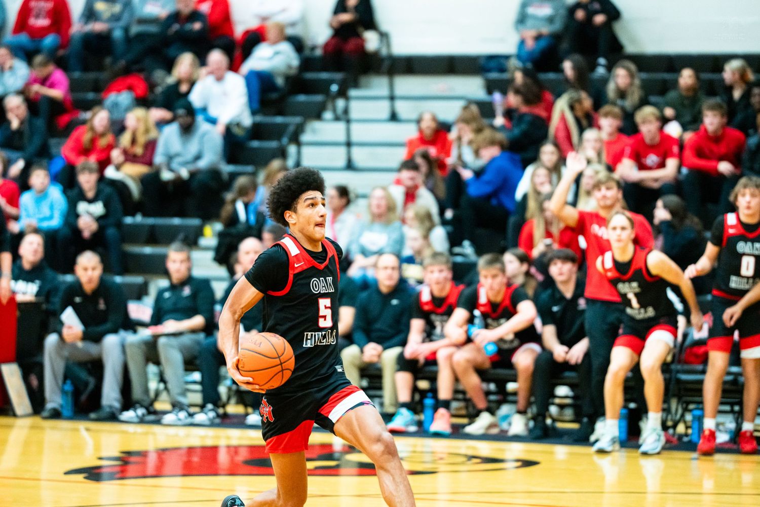 Energetic high school basketball player number 5 in red uniform dribbles the ball during a game with fans watching.