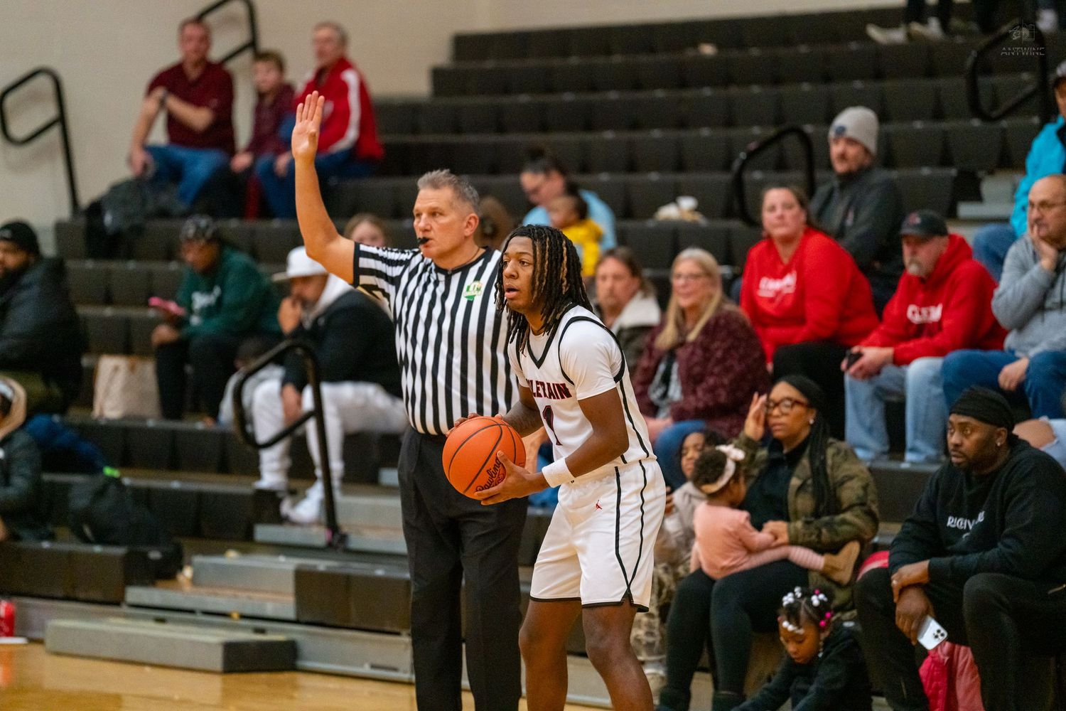 High school basketball player holds the ball near the sideline as a referee signals during a game.