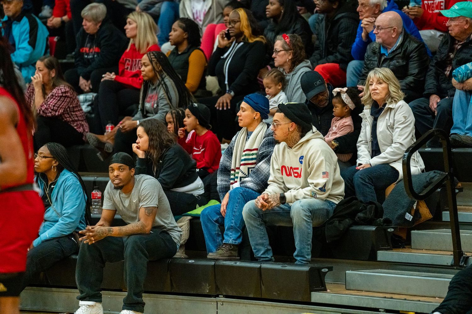 Spectators sitting courtside on bleachers watching a high school basketball game in a red and white gymnasium.