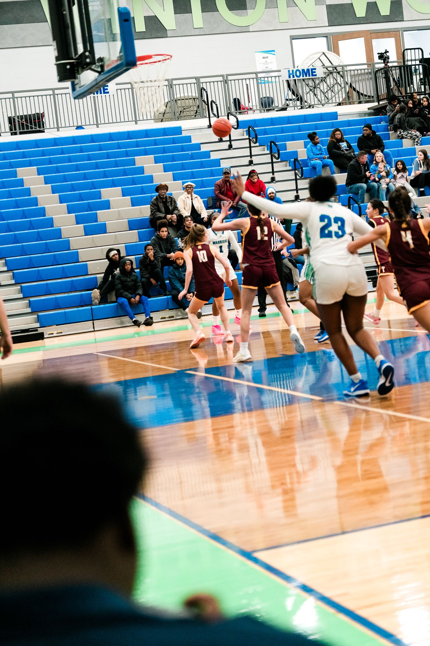 High school girls basketball players battle for the ball near the basket during a game in a blue gymnasium.