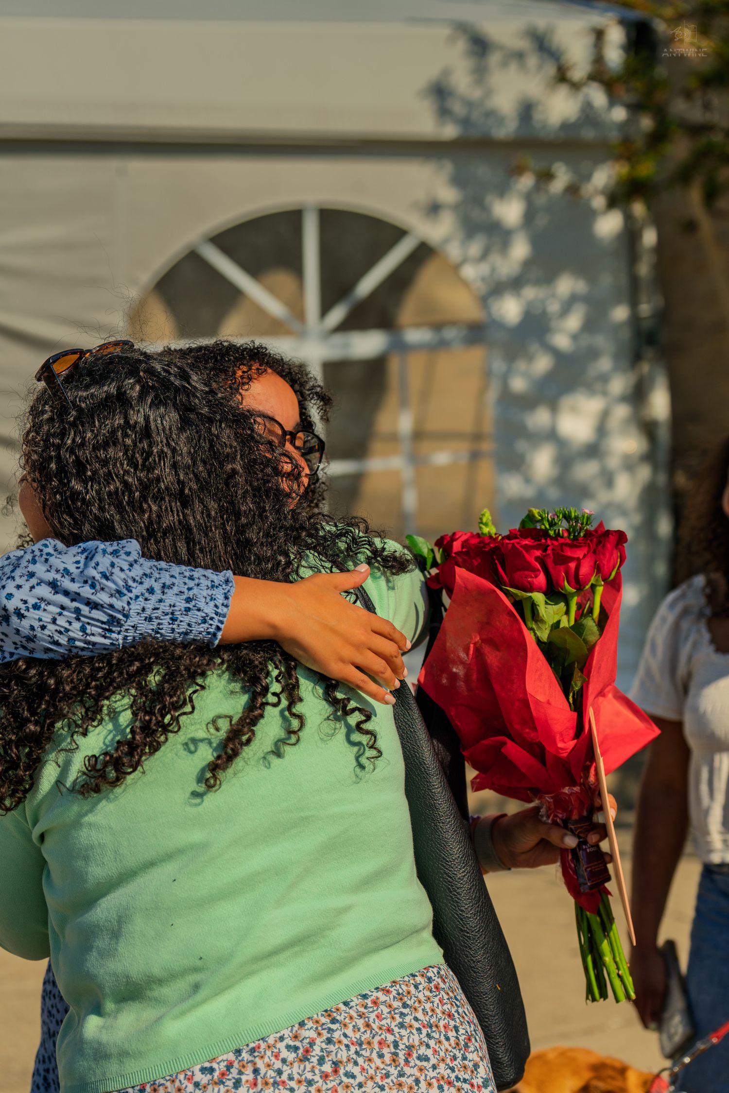 Two women hug warmly outdoors, one holding a vibrant bouquet of red roses in sunlight.