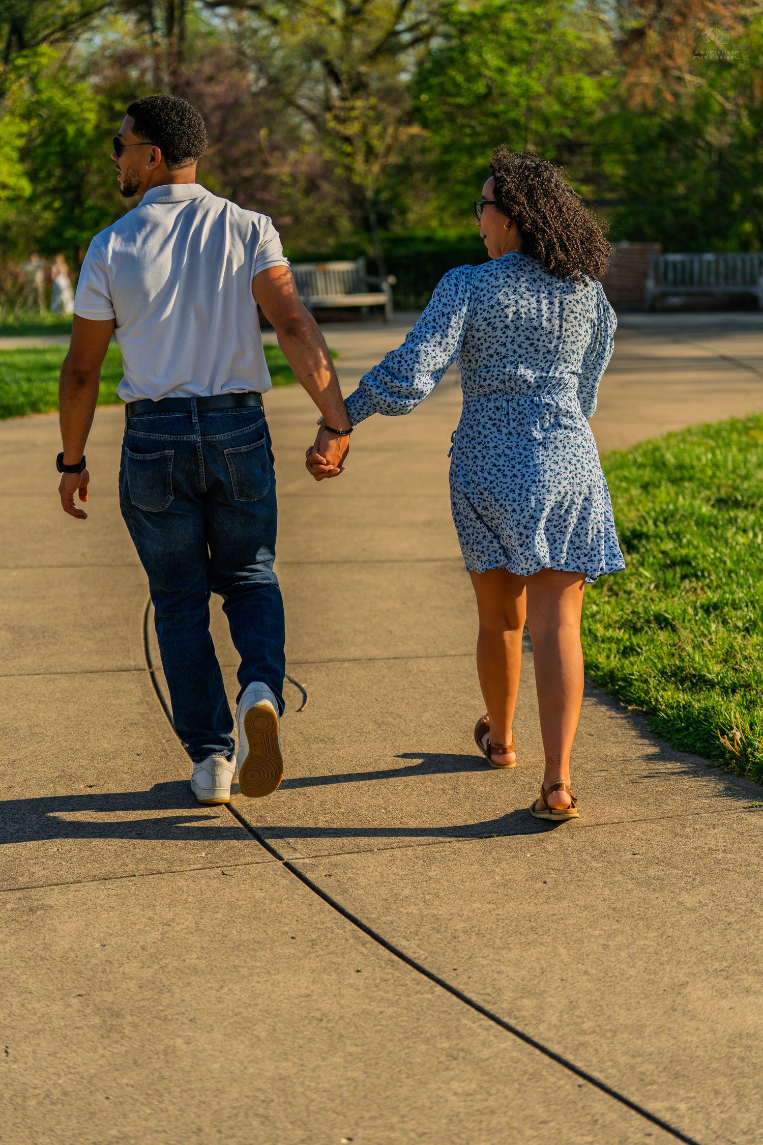 Couple walking hand in hand along a sunlit park pathway, woman in blue floral dress looks back.