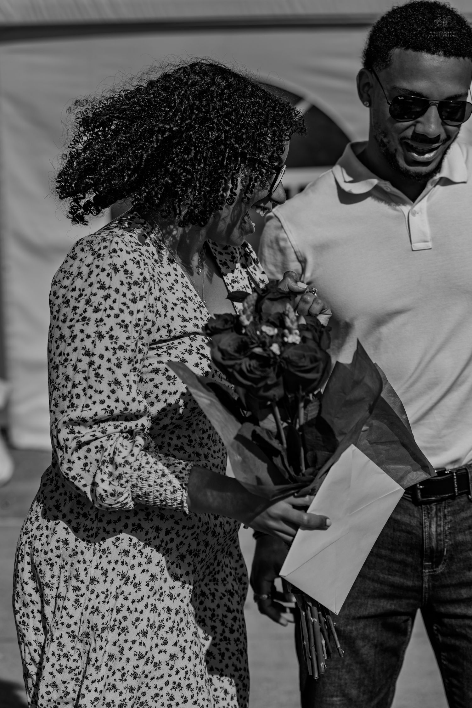 Black and white image of a man presenting roses and a card to a woman in a floral dress outdoors.