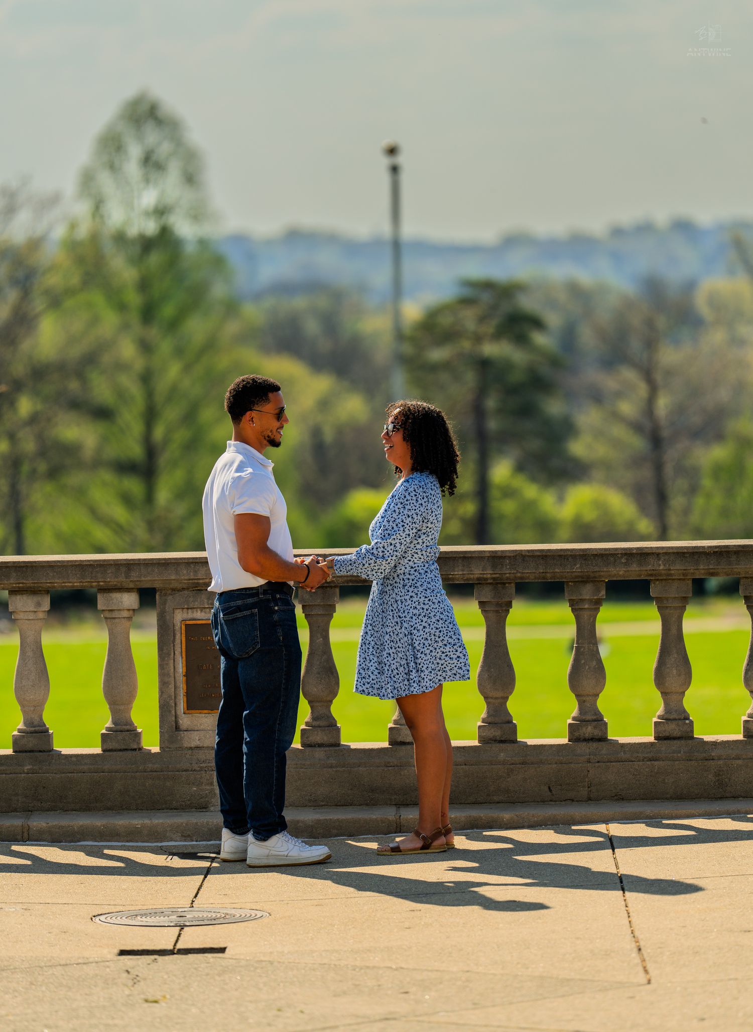 Couple facing each other holding hands on a sunny stone terrace with green landscape behind.