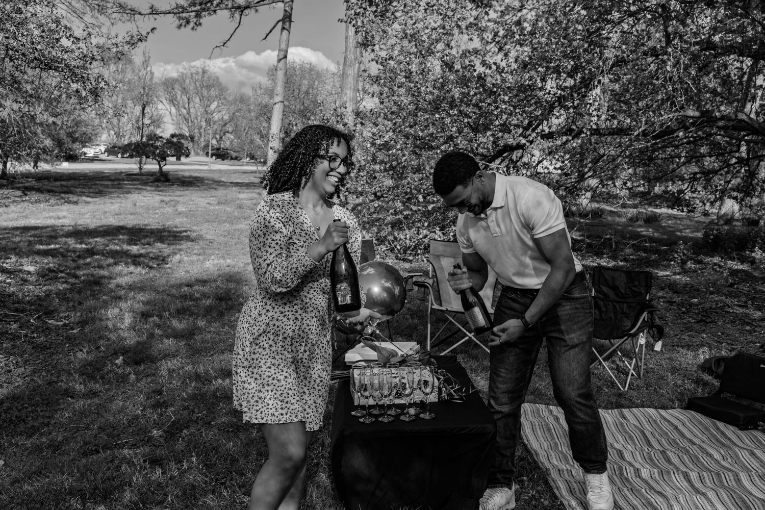 Black and white image of couple at outdoor picnic table with champagne bottle and decorative setup.