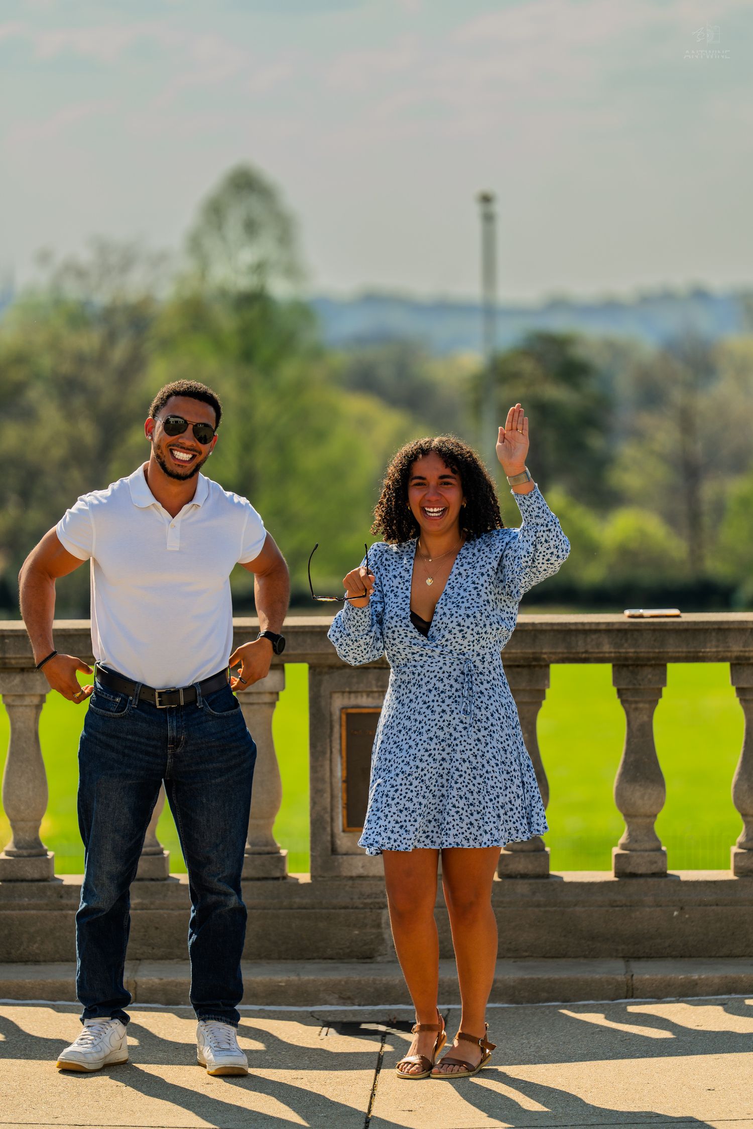 A smiling couple stands on a sunlit stone balcony terrace, woman waving with park views behind.