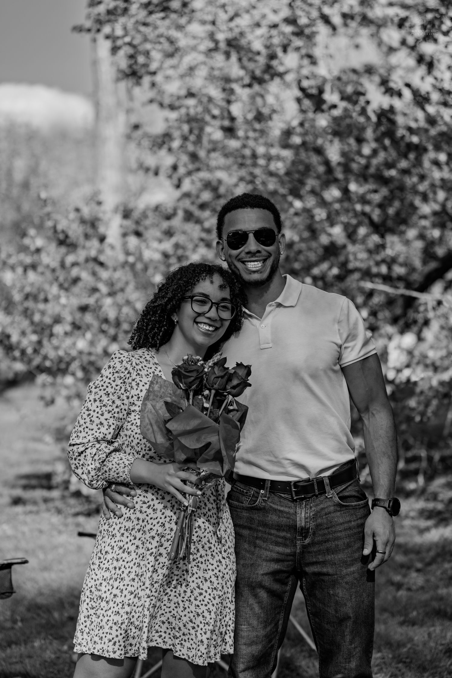 Black and white portrait of smiling couple holding roses bouquet in blooming orchard garden.