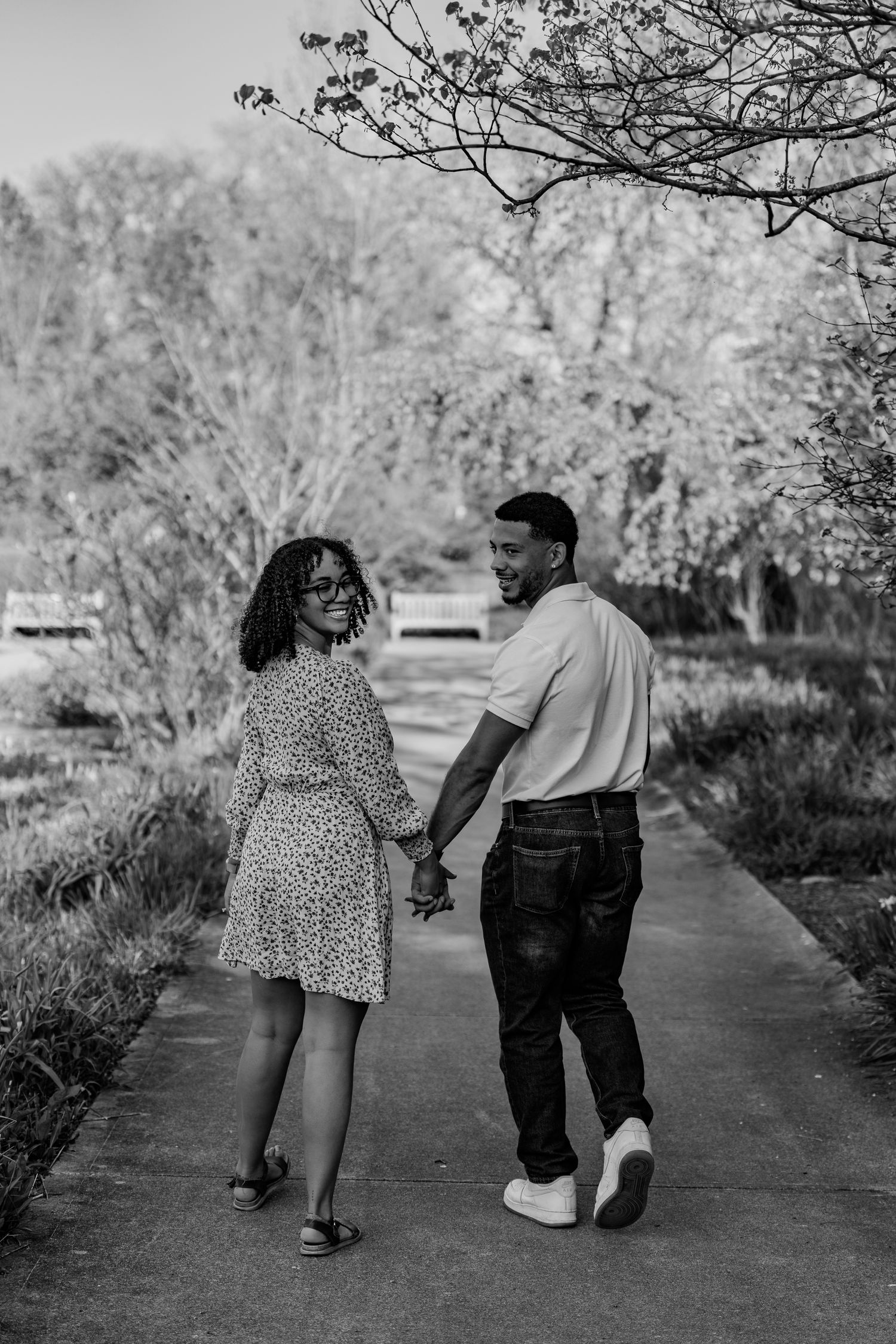 Black and white photo of a couple holding hands and walking on a park path, woman in floral dress looking back.