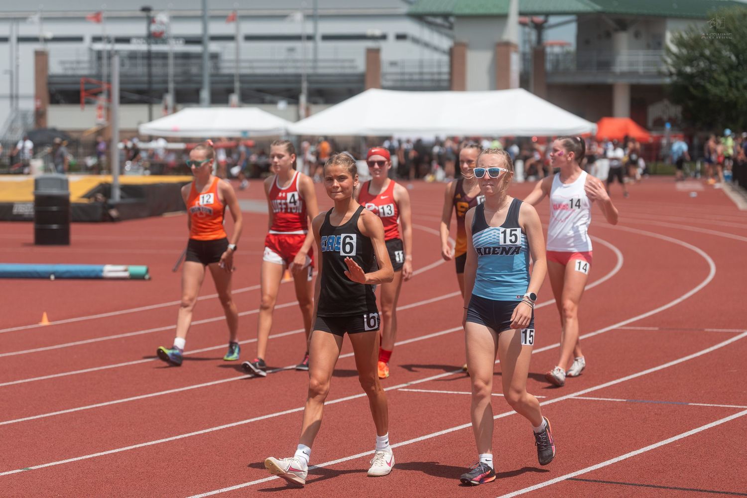 Group of female runners in numbered bibs competing in outdoor track race on sunny day at stadium.