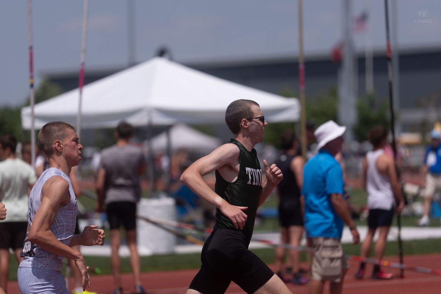 A young runner wearing glasses competes in a track and field event with white tents visible in the background.