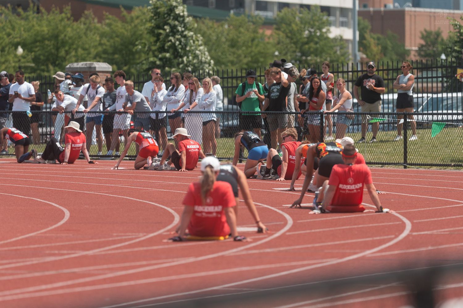Athletes in starting positions on red track at outdoor collegiate track and field competition with crowd watching.