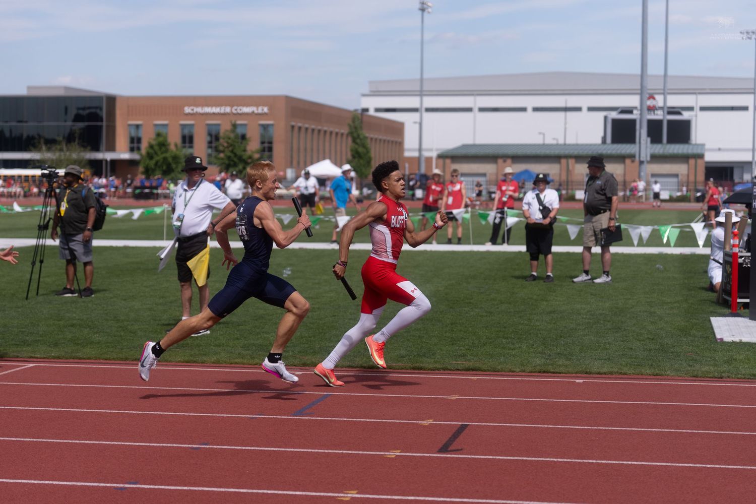 Two athletes sprint side by side on red track at outdoor track and field competition on sunny day.