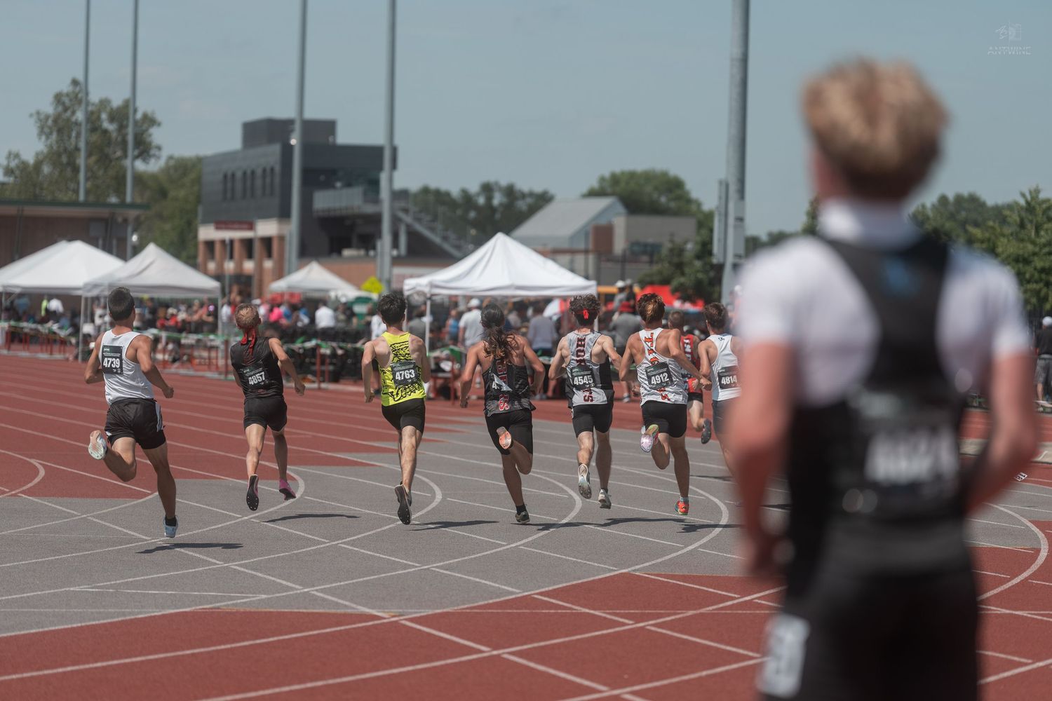 Runners compete in a track race at an outdoor stadium during a sunny day track and field meet.