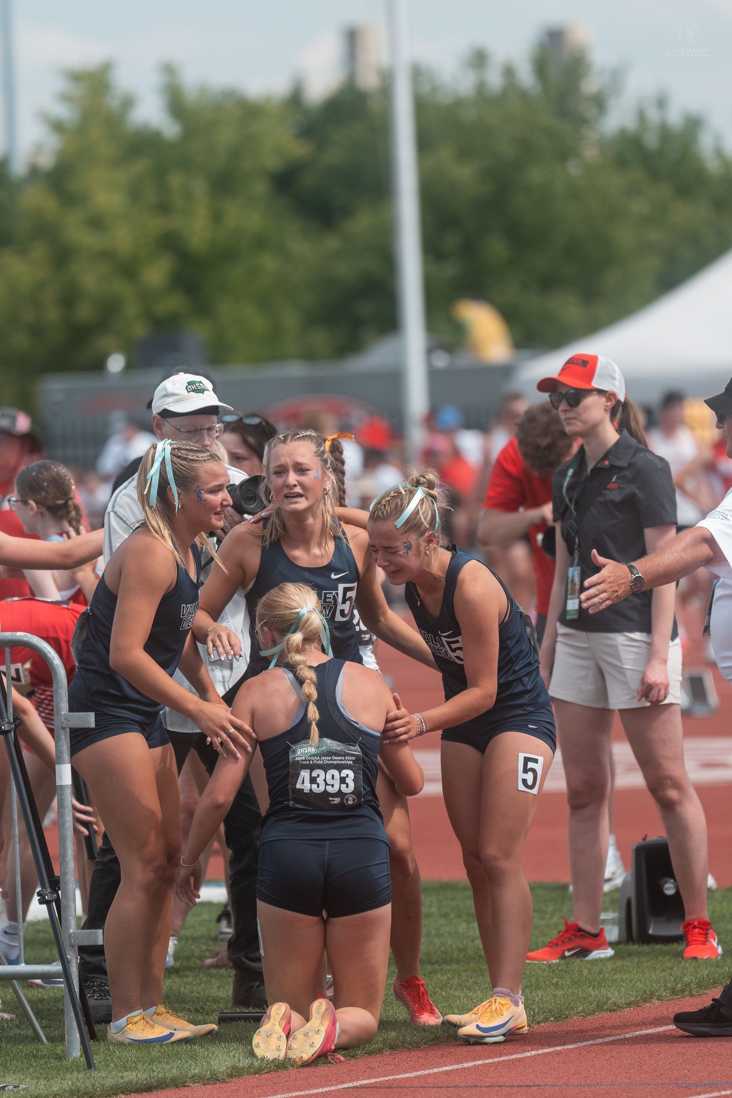 Female track team in navy uniforms emotionally supports a kneeling teammate wearing bib 4393 after a relay race.