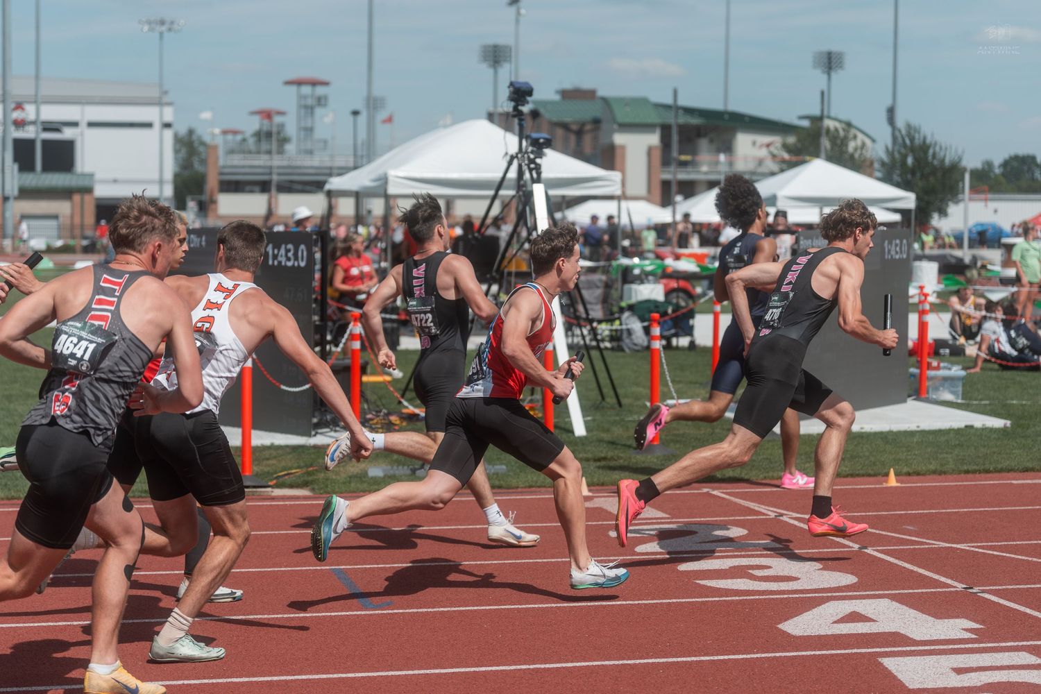 Multiple relay teams execute baton exchanges simultaneously on red track at high school track championship meet.