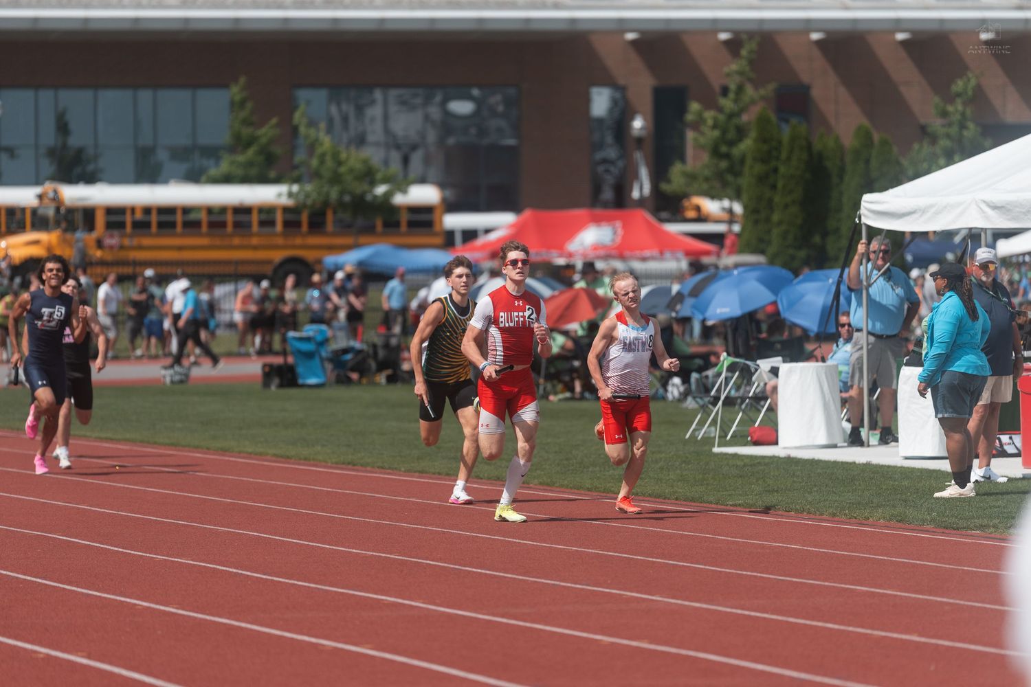 Track athletes race past spectators and white tents during an outdoor high school track and field competition.