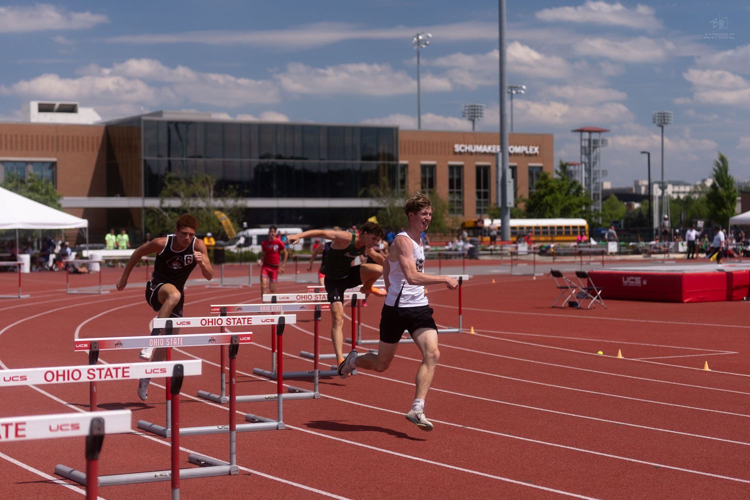 High school athletes clear hurdles on the Ohio State University red track during a sunny championship meet.