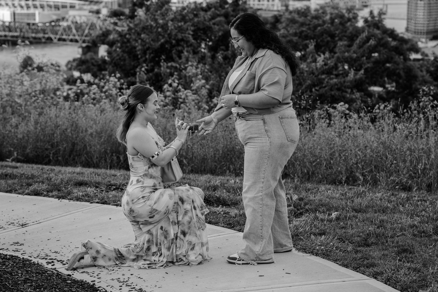 Woman kneeling on path proposing to standing partner in black and white photo.