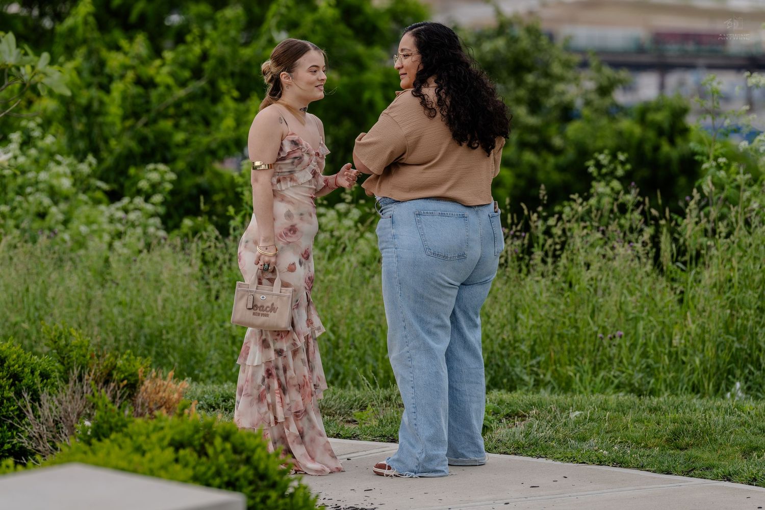 Two women stand on a garden path, one in a floral dress and one in casual jeans, engaged in conversation.