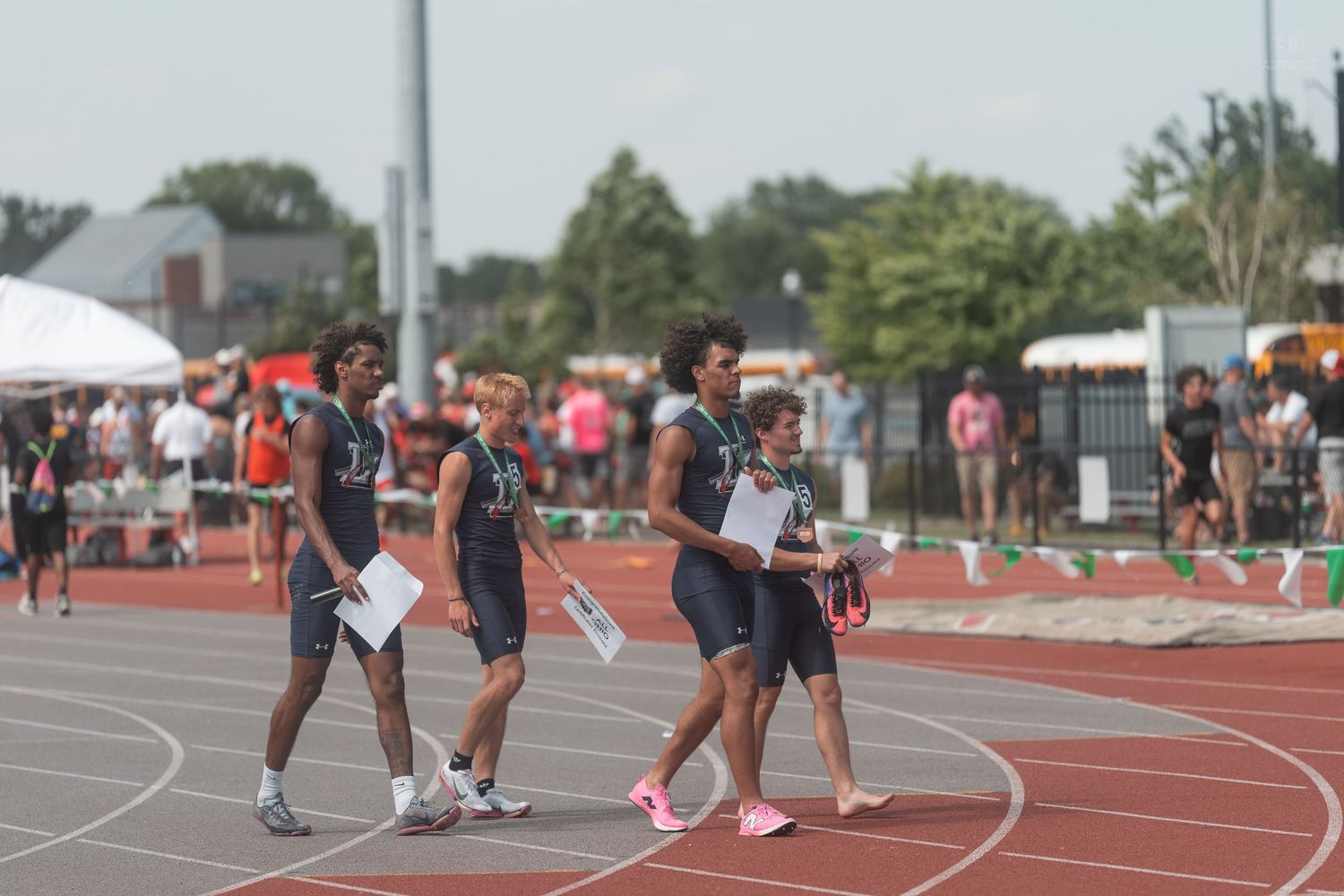 Track athletes in dark uniforms walk along a red running track during an outdoor track and field competition.