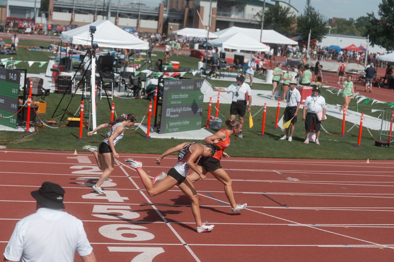 Runners sprint toward finish line at outdoor track and field competition with white tents and crowd in background.