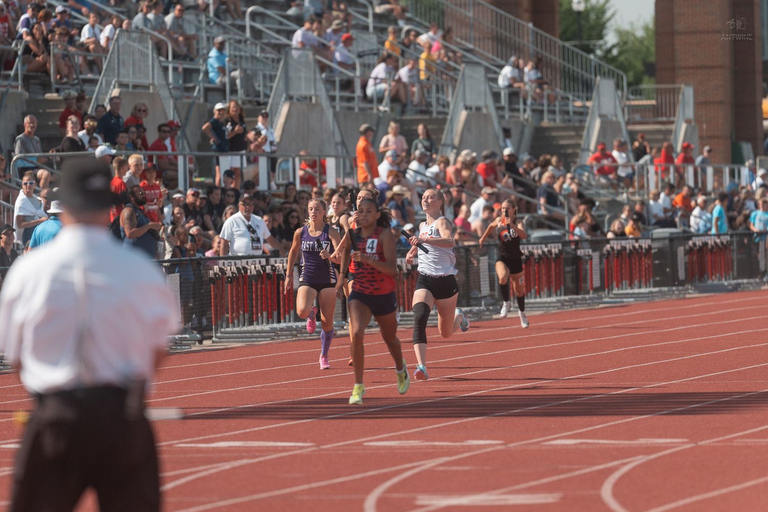 Female runners race down a red track in front of packed stadium bleachers at a track meet.