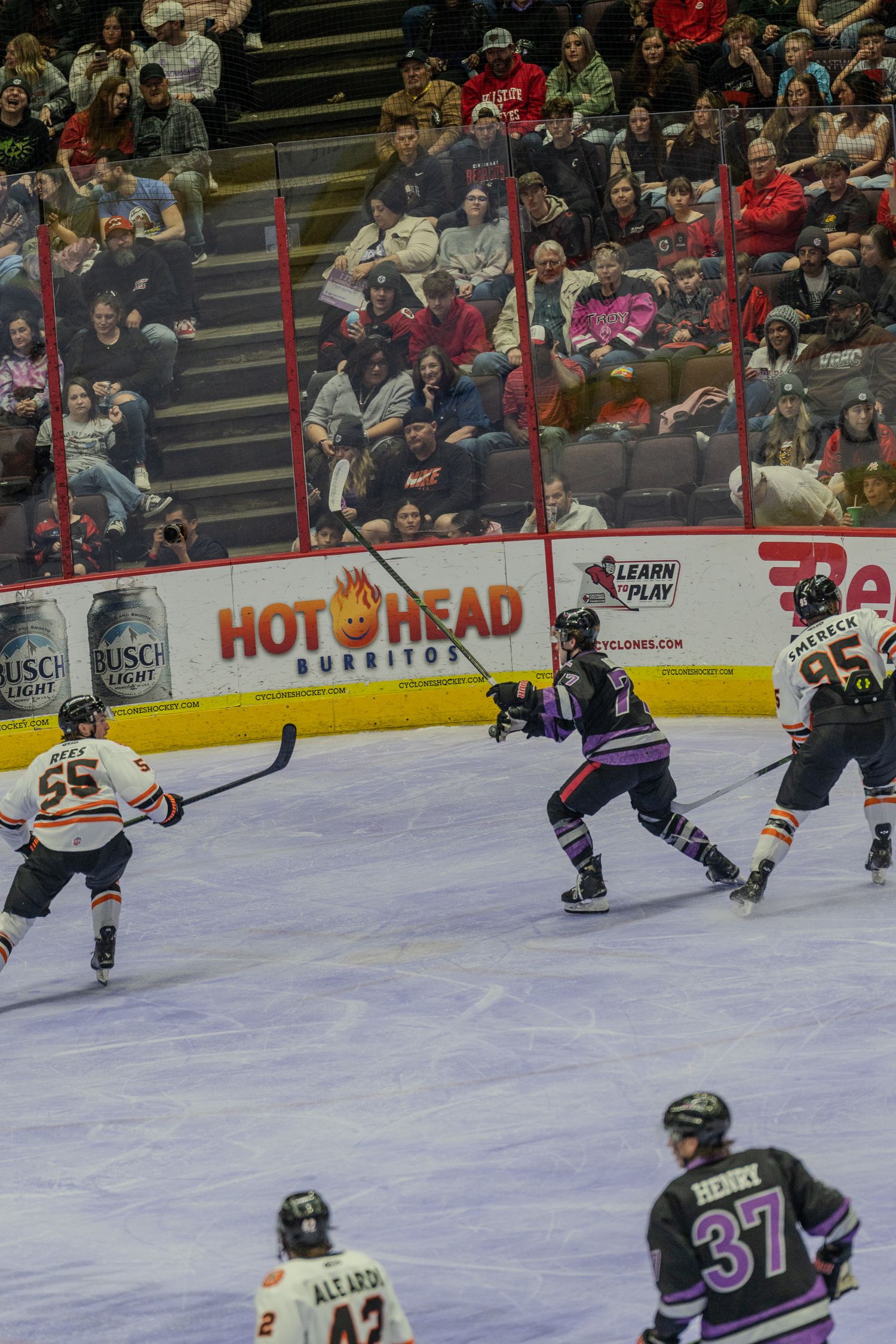 Ice hockey players in orange and purple uniforms clash near the boards with fans watching from stands.