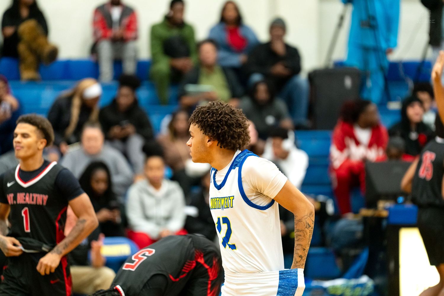 High school basketball player #12 in blue uniform stands on court during game against red-uniformed opponents.