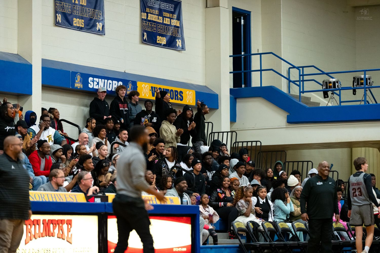 Large crowd fills gymnasium bleachers watching high school basketball game with blue and yellow decor.