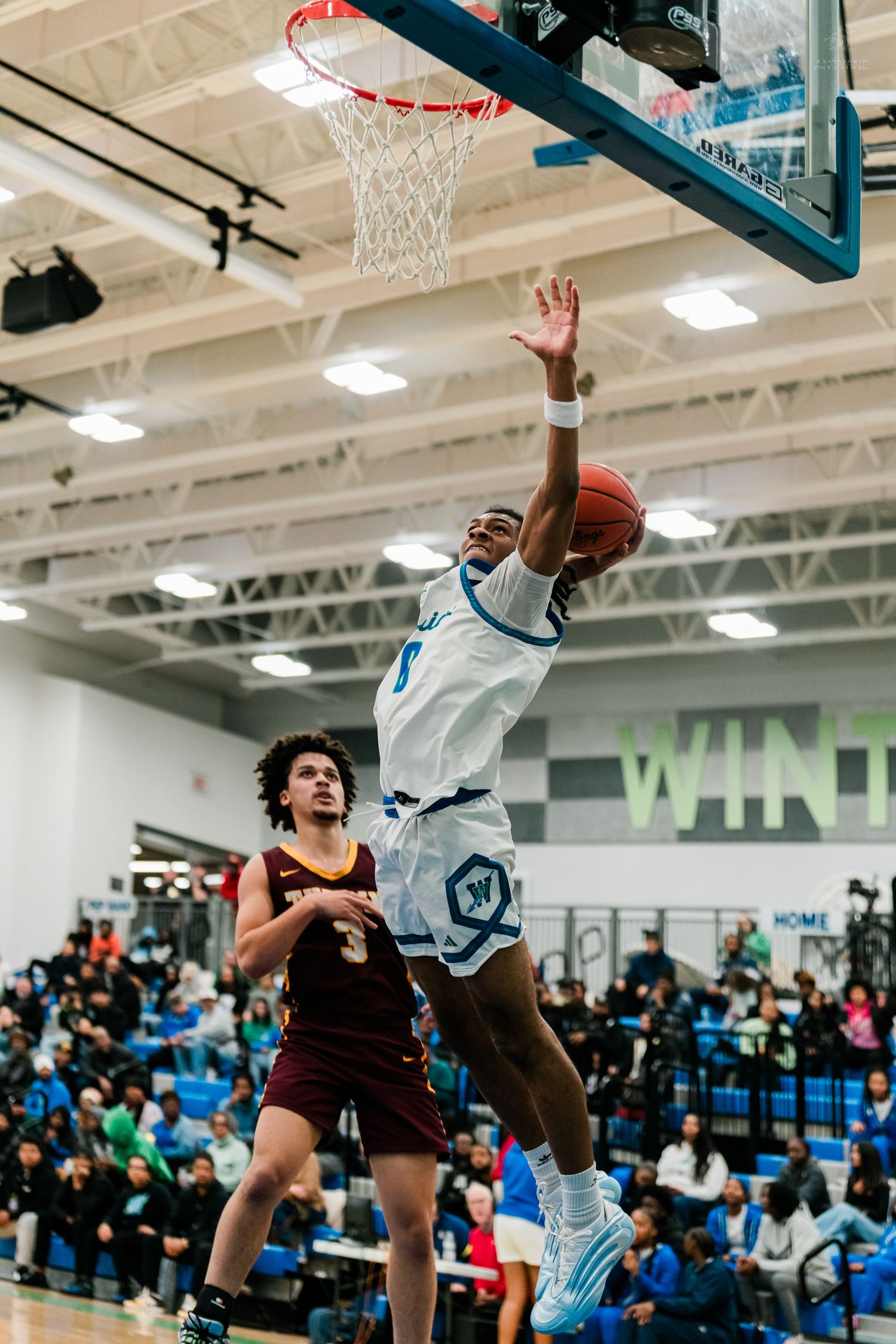 A player in white leaps for a layup while a maroon #3 defender watches during a high school basketball game.