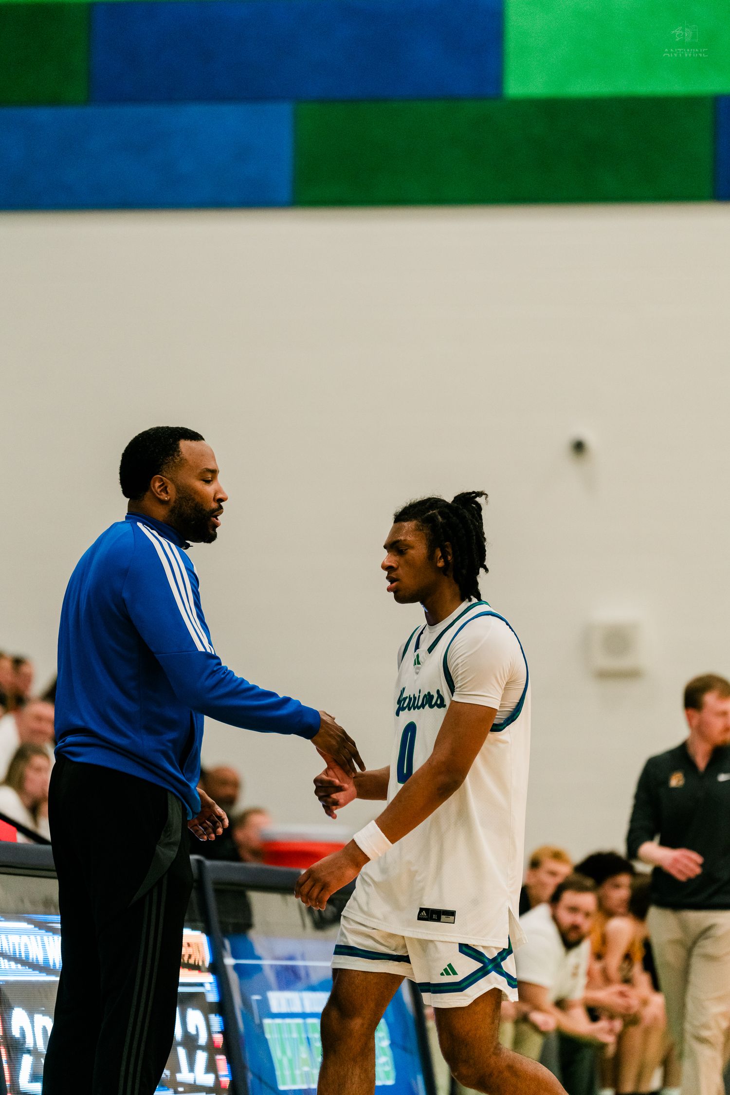 Basketball coach in blue Adidas jacket shakes hands with player number 0 on the sideline.