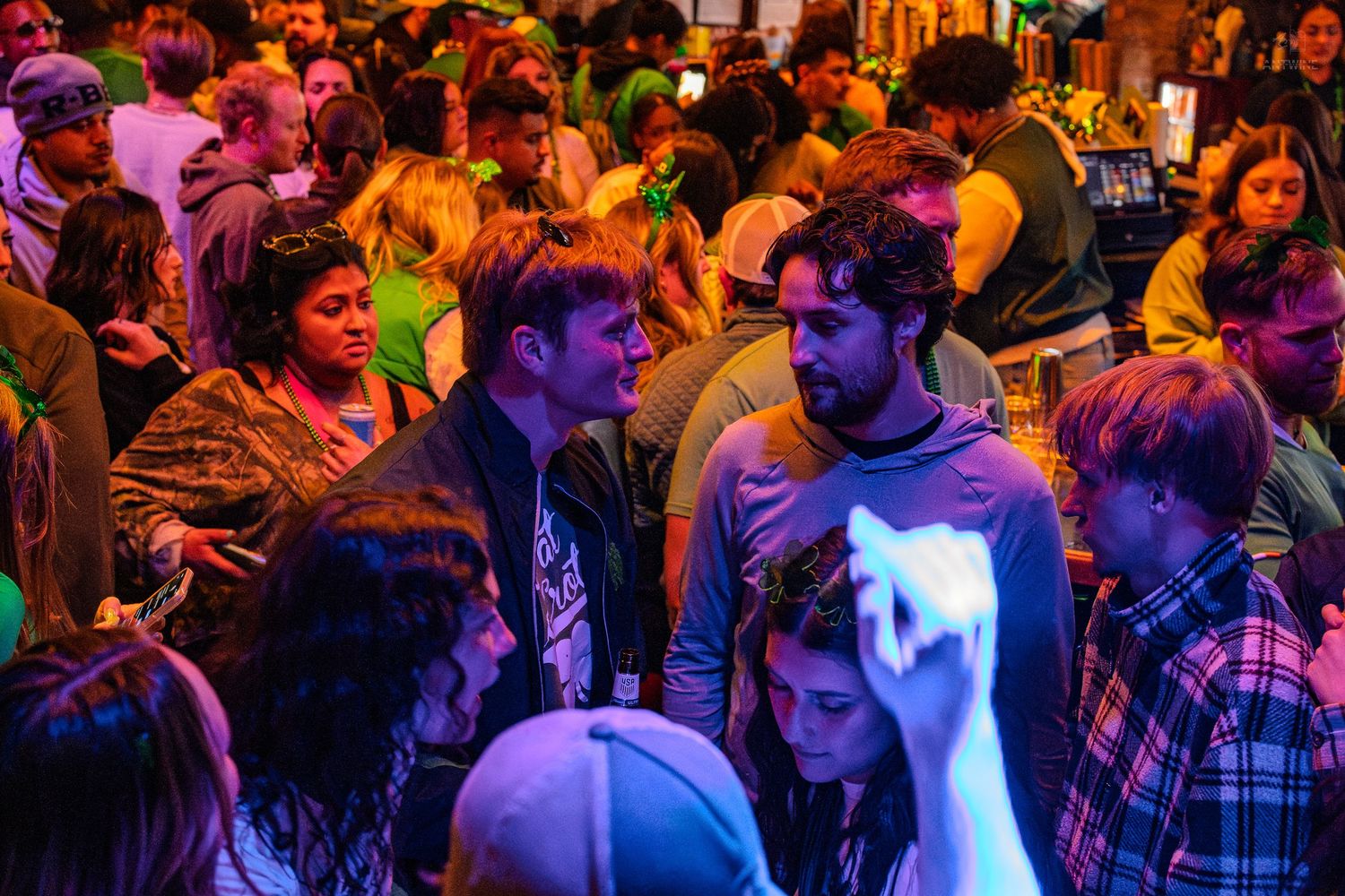 Diverse crowd of young people mingling under warm orange and green lighting at a St. Patrick's Day event.