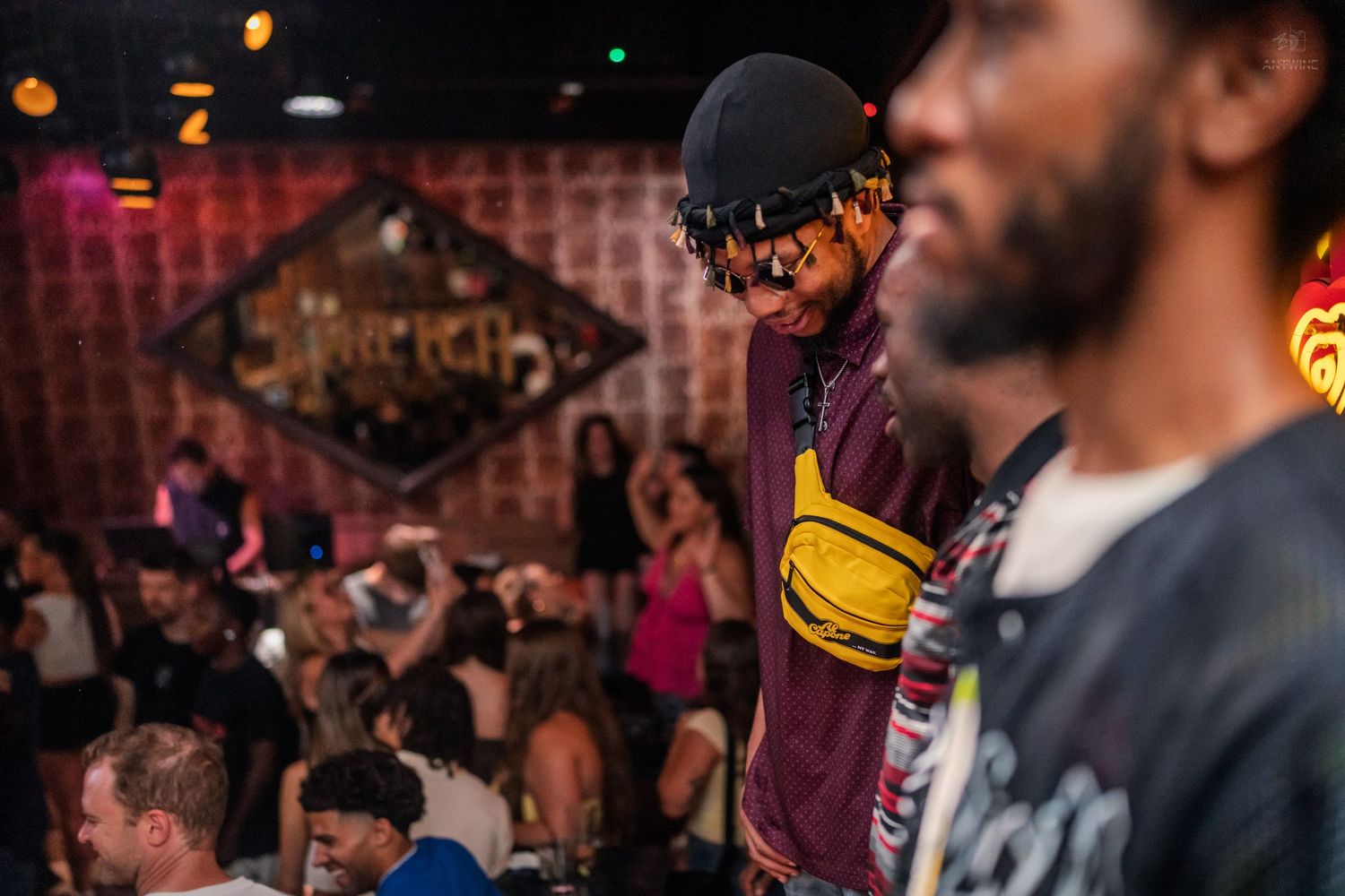 A man wearing a cap and yellow fanny pack performs at a packed nightclub with colorful decor.