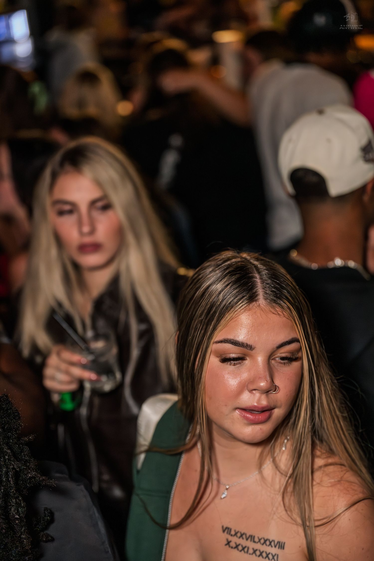 Two young women at a nightclub, one blonde holding a cocktail glass in a crowded venue.