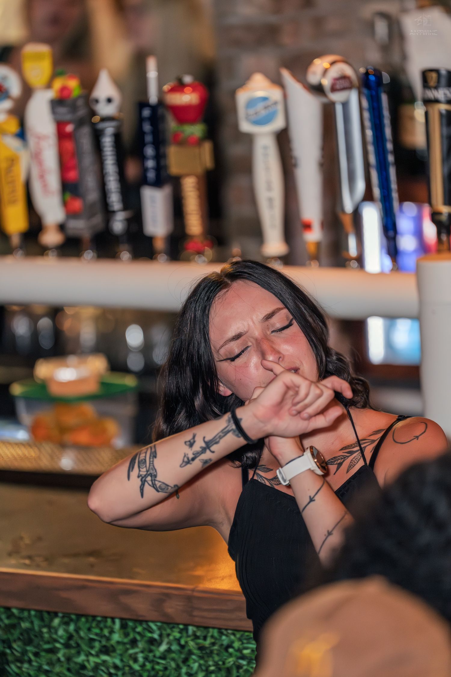 Tattooed woman in black top leaning on bar counter with draft beer taps in background.