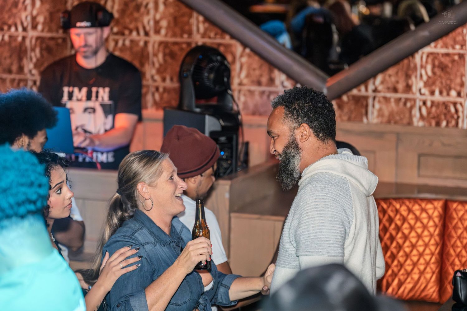 Two people engaged in conversation at a lively nightclub with a DJ in the background.