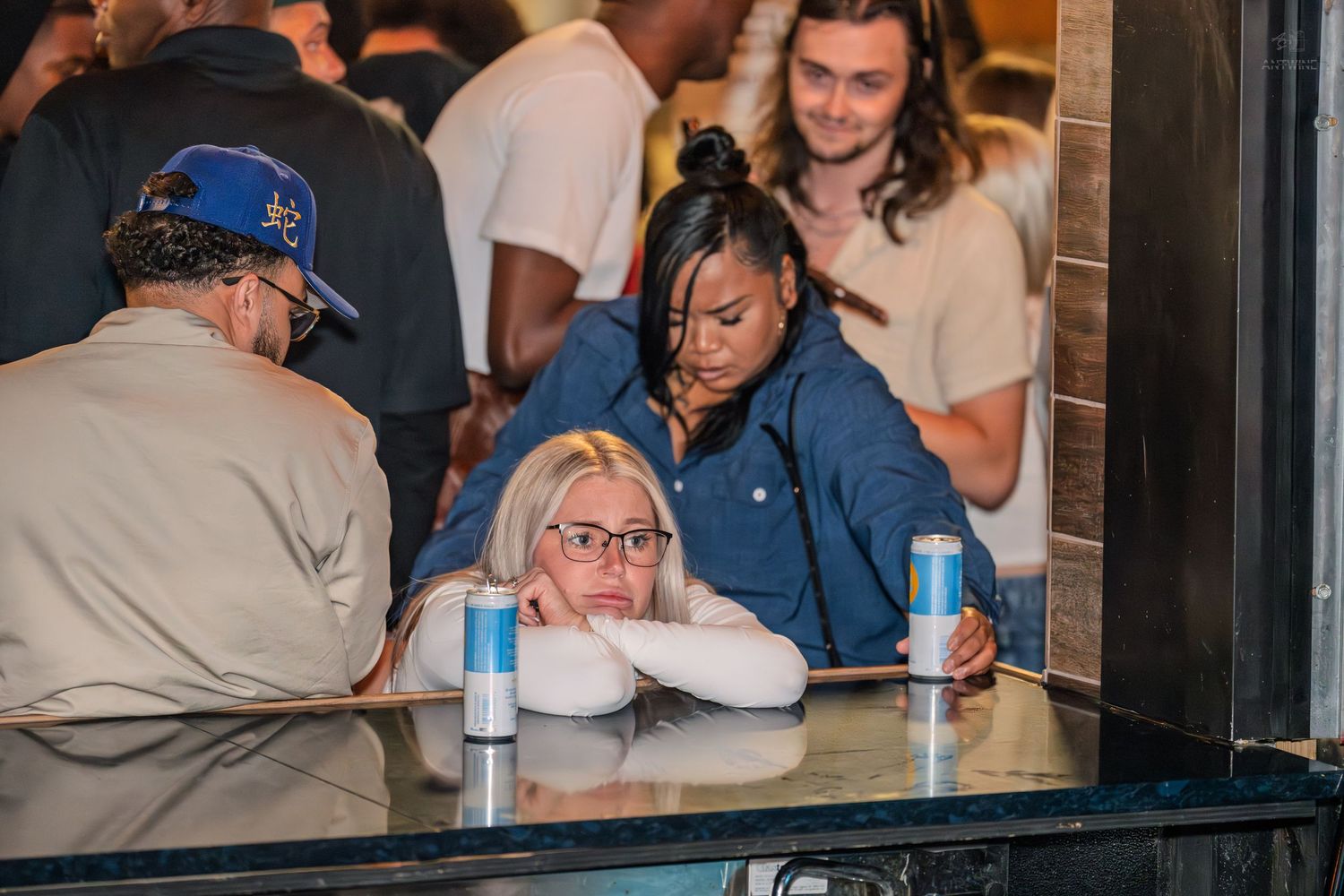 Two women sitting at a bar table with canned drinks, one resting her head on her arms looking tired.